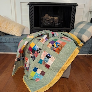 May include: A patchwork quilt featuring a light green fabric with a small white floral pattern as the primary background. The quilt is composed of various square blocks containing multi-colored fabric scraps and has a bright yellow border.