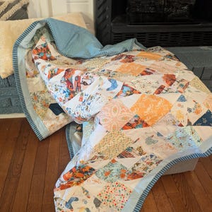 May include: A handmade quilt with a patchwork design featuring various patterns and colors, including orange, blue, and floral prints. The quilt has a light blue border and is draped over a surface.