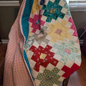 May include: A patchwork quilt featuring a variety of colorful fabric squares in shades of red, yellow, blue, and pink. The quilt is constructed with a grid pattern of small squares and includes floral, polka dot, and gingham prints, with a light pink gingham backing.