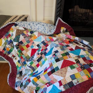 May include: A handmade patchwork quilt with a vibrant mix of colors and patterns. The quilt features squares and triangles in various fabrics, including floral, geometric, and solid colors. It has a burgundy border and is draped over a surface.