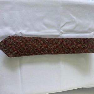 Peut inclure: Une cravate en tissu tissé texturé avec un motif à carreaux diagonaux rouge foncé et vert olive. La cravate présente une extrémité pointue et une largeur constante sur toute sa longueur, mettant en valeur une construction dense en fils.