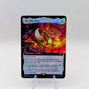 May include: A holographic trading card titled The One Ring, featuring a golden ring centered against a swirling background of red, orange, and black flames. The card text includes Legendary Artifact, Indestructible, and game rules.