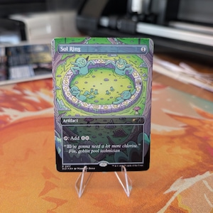 Peut inclure: Une carte à collectionner rectangulaire intitulée Sol Ring avec une illustration verte de gobelins dans une piscine circulaire. Le texte de la carte inclut Artifact, le symbole de mana et la citation : We're gonna need a lot more chlorine.