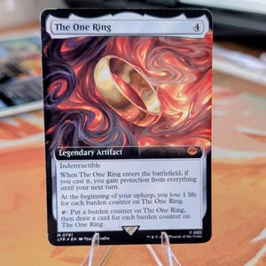 Peut inclure: Une carte à collectionner rectangulaire intitulée The One Ring, représentant un anneau doré centré sur un fond tourbillonnant de fumée rouge, orange et noire. Le texte de la carte inclut Legendary Artifact, Indestructible et les règles.