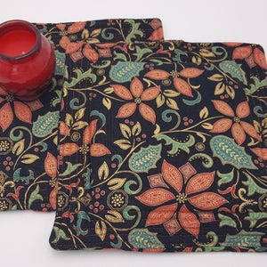 Puede incluir: Dos posavasos de tela negra con un patrón floral dorado, rojo y verde. Los posavasos son cuadrados y tienen un borde cosido.
