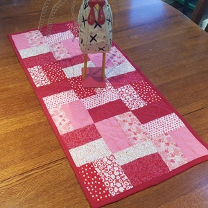 May include: A red and pink quilted table runner with a white heart pattern. The runner is made of fabric squares sewn together in a patchwork design.