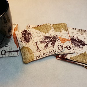 May include: Set of six fabric coasters with a vintage autumn theme. The coasters feature a cream background with brown, orange, and green accents. The word "AUTUMN" is printed on each coaster.
