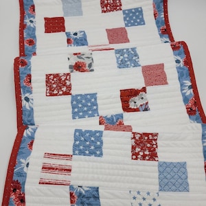 Puede incluir: Un camino de mesa blanco con un diseño de patchwork rojo, blanco y azul. El camino de mesa está acolchado y tiene un borde rojo con lunares blancos.