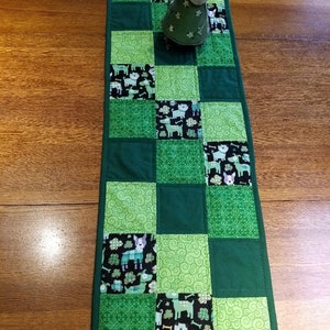 May include: A green and black patchwork table runner with a dog and shamrock pattern. The runner is made of fabric squares sewn together.