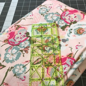 May include: A folded floral fabric with pink, blue, and white flowers is shown with a clear ruler. The ruler is marked with measurements in inches. The fabric is on a black and gray cutting mat.
