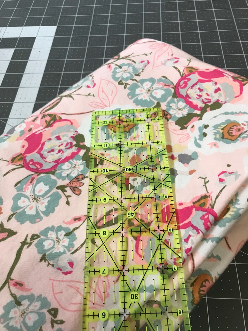 May include: A folded floral fabric with pink, blue, and white flowers is shown with a clear ruler. The ruler is marked with measurements in inches. The fabric is on a black and gray cutting mat.