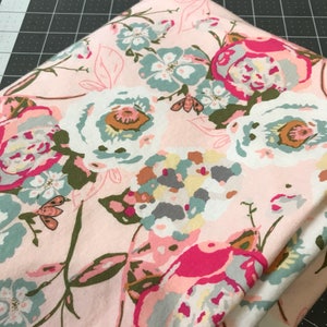 May include: Folded fabric featuring a floral pattern in pink, white, teal, and fuchsia. The design includes large blooms, leaves, and small insect illustrations. The fabric is folded over, showing the pattern's repeat.