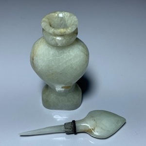 Puede incluir: Pequeño frasco de perfume de piedra verde claro con tapón a juego. El frasco tiene un cuerpo redondeado y una parte superior acampanada. El tapón es largo y delgado, con un extremo redondeado. El frasco y el tapón son de piedra translúcida.