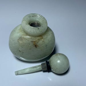 Peut inclure: Une bouteille en pierre vert clair avec un corps rond et un dessus circulaire. La bouteille a un bouchon assorti avec une petite poignée ronde. Le bouchon est détaché, révélant une pièce métallique filetée.