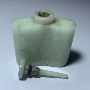 May include: A pale green, rectangular perfume bottle with a cylindrical neck and a detached stopper. The bottle has a slightly rounded top and visible cracks. The stopper has a black ring and a threaded metal section.