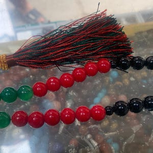 May include: A beaded strand with green, red, and black beads. The beads are strung together, forming a loop. A tassel with red and green threads is attached to the top. The beads are likely made of polished stone.
