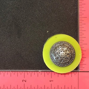 Könnte beinhalten: Ein silberfarbener Metallknopf mit einem dekorativen Design, eingebettet in eine gelbe Harzform.