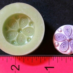 Könnte beinhalten: Eine Silikonform zum Herstellen eines dekorativen Knopfes mit Blumenmuster. Die Form ist hellgrün und der Knopf ist lila.
