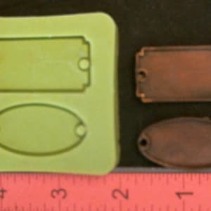 Puede incluir: Un molde de silicona verde claro con cavidades rectangulares y ovaladas, junto a dos etiquetas de metal bronce. Una regla muestra las medidas en centímetros. El molde es para crear etiquetas.