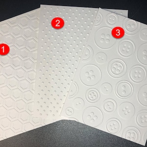 May include: Three white textured embossing folders. The first folder has a hexagonal pattern. The second folder has a small dot pattern. The third folder has a button pattern.
