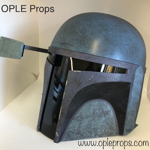 May include: A blue and gray Mandalorian helmet with a weathered finish. The helmet has a black visor and a small, rectangular attachment on the side. The text "www.opleprops.com" is visible at the bottom of the image.