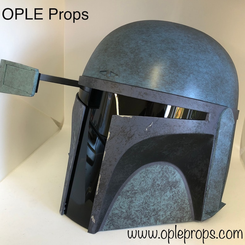 May include: A blue and gray Mandalorian helmet with a weathered finish. The helmet has a black visor and a small, rectangular attachment on the side. The text "www.opleprops.com" is visible at the bottom of the image.