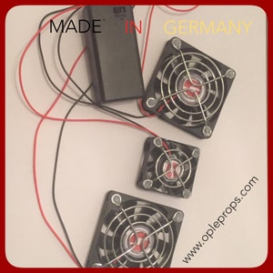 May include: Three black computer fans with metal grills and red wires. The fans are connected to a black battery pack. The text "MADE IN GERMANY" is visible at the top of the image. The text "www.oppleprops.com" is visible at the bottom right of the image.