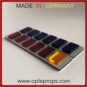 May include: A close-up of a silver panel with a row of colored buttons. The buttons are red, orange, yellow, green, blue, and purple. The panel is labeled "MADE IN GERMANY" and "www.opleprops.com".