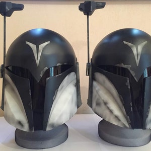 May include: Two black and white Mandalorian helmets with a white V symbol on the front. The helmets are on display stands.