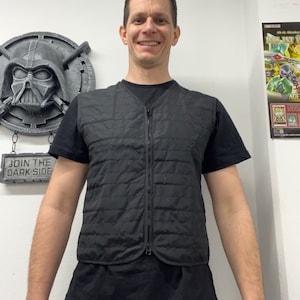 May include: A black quilted vest with a full-length zipper is worn over a black t-shirt. The vest has a diamond pattern. The person is wearing blue jeans and a watch. A Star Wars themed wall decoration is visible in the background.