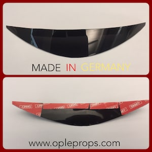 May include: Black plastic car part with adhesive tape on the back. The part is curved and has a smooth surface. The text "MADE IN GERMANY" is printed on the white background below the part. The text "www.opleprops.com" is printed on the white background below the part.