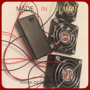 May include: Three small black computer fans with silver wire mesh grilles and red and black wires. The fans are connected to a black battery pack labeled "OP-0204" and the text "MADE IN GERMANY" is visible above the fans.