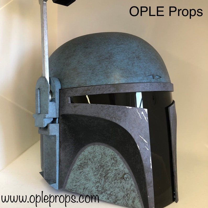 May include: A blue and black Mandalorian helmet with a weathered finish. The helmet has a visor and a rangefinder on top. The helmet is made by OPLE Props and the website is www.opleprops.com.