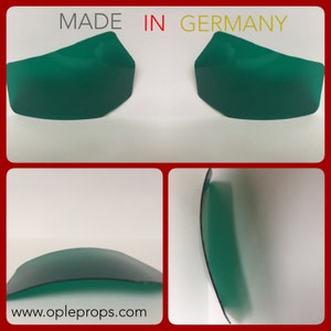 Puede incluir: Dos piezas de plástico verde curvadas, una encima de la otra. La pieza superior es ligeramente más grande que la pieza inferior. Las piezas están hechas en Alemania. El texto "MADE IN GERMANY" está impreso en rojo y amarillo sobre un fondo blanco.