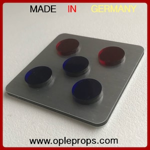 Könnte beinhalten: Eine rechteckige, gebürstete Metallplatte mit fünf kreisförmigen, farbigen Teilen. Zwei rote und drei blaue Teile sind auf der Platte angeordnet. Der Text "MADE IN GERMANY" und die Website-Adresse "www.opleprops.com" sind sichtbar.