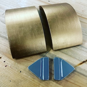 Könnte beinhalten: Zwei goldfarbene, gebogene Metallstücke und zwei graue, dreieckige Teile sind auf einer Holzoberfläche angeordnet. Die goldenen Teile haben eine gebürstete Textur. Die grauen Teile sind glänzend und haben die Form rechtwinkliger Dreiecke.