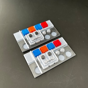 Può includere: Due pannelli di controllo stampati in 3D con pulsanti e manopole. I pannelli sono grigi con accenti bianchi. I pulsanti sono rossi, arancioni, blu e neri. Le manopole sono argentate.