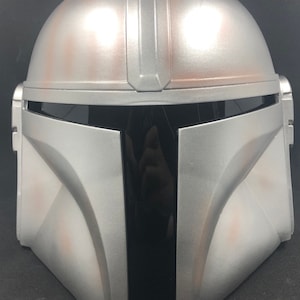 May include: Silver Mandalorian helmet with a black visor. The helmet has a weathered appearance with brown accents and the text "OPLE Props" and "www.opleprops.com".