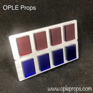 May include: A silver metal plaque with the text "OPLE Props" at the top. The plaque has eight rectangular, colored glass inserts. The top row has four red inserts and the bottom row has four blue inserts. The text "www.opleprops.com" is at the bottom of the plaque.