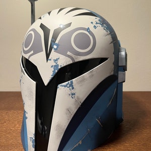 May include: A Mandalorian helmet with a white, blue, and gray color scheme. The helmet features a distinctive design with a visor and a rangefinder. The helmet has a weathered appearance, with distressed paint details.