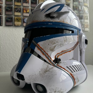 May include: A white and blue helmet with a weathered appearance, featuring blue accents and orange detailing. The helmet has a visor and speaker grilles, and is likely a costume or collectible item.