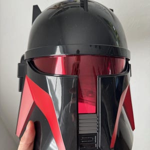 May include: Black and red helmet with a visor. The helmet has a glossy finish and features red accents on the sides and front. A black antenna-like structure extends from the top. The visor is a deep red color.