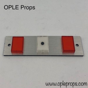 Puede incluir: Un panel de metal plateado con tres botones. Los botones son rectangulares y tienen una cubierta de pl&aacute;stico transparente. Los botones izquierdo y derecho son rojos, y el bot&oacute;n central es blanco con un punto negro en el centro. El panel tiene el texto "OPLE Props" en la parte superior y "www.opleprops.com" en la parte inferior.