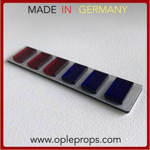Op de afbeelding: Een zilveren metalen balk met zeven rechthoekige rode en blauwe glazen inzetstukken. De balk heeft de tekst "MADE IN GERMANY" bovenaan en "www.opleprops.com" onderaan.