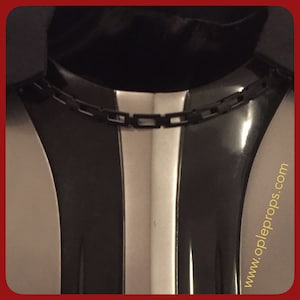 Könnte beinhalten: Nahaufnahme eines schwarz-silbernen Kostümteils mit Kettendetail. Das Design zeigt eine zentrale silberne Platte, flankiert von schwarzen, gebogenen Abschnitten. Die Website www.opleprops.com ist unten rechts sichtbar.