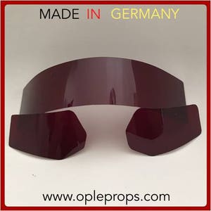 May include: Three pieces of dark red plastic, likely for a costume or prop. The top piece is a curved visor, and the two bottom pieces are shaped like eyes. The text "MADE IN GERMANY" is visible at the top of the image.