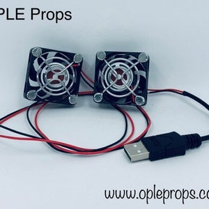 Könnte beinhalten: Zwei kleine schwarze Computerlüfter mit silbernen Gittern und roten Kabeln, die mit einem schwarzen USB-Kabel verbunden sind. Die Lüfter sind mit "OPLE Props" beschriftet und die Website "www.opleprops.com" ist unter den Lüftern aufgedruckt.