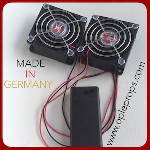 May include: Two small black fans with chrome grills and red wires connected to a black battery pack. The fans are labeled "MADE IN GERMANY" and the battery pack has the website "www.opleprops.com" printed on it.