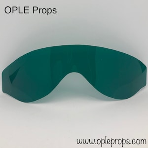 May include: A single, curved, dark green lens, likely for eyewear or a prop. The lens has a smooth, reflective surface and a distinctive shape, with a curved top and a dip in the center. The text "OPLE Props" and "www.opleprops.com" are visible.