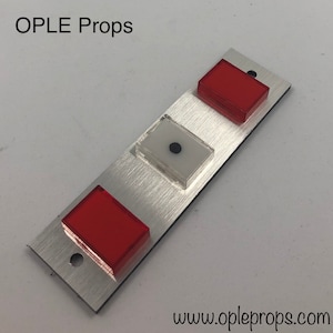 Puede incluir: Un panel de metal plateado con tres botones. El bot&oacute;n superior es rojo, el bot&oacute;n del medio es transparente con un punto negro y el bot&oacute;n inferior es rojo. El panel tiene el texto "OPLE Props" y "www.opleprops.com" impreso en &eacute;l.
